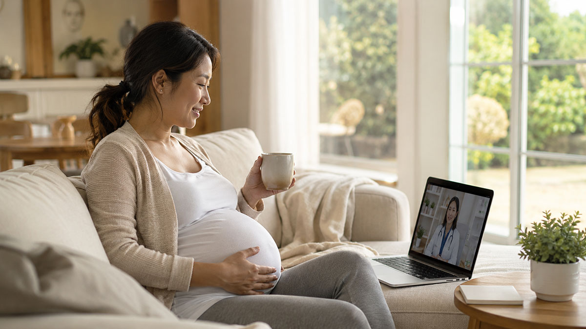 A pregnant Asian woman, wearing a beige cardigan and white top, sitting comfortably on a grey sofa in a sunlit living room and having a telehealth video call on her laptop. She holds a mug in one hand and gently rests her other hand on her pregnant belly, looking toward the laptop screen which displays a female doctor.